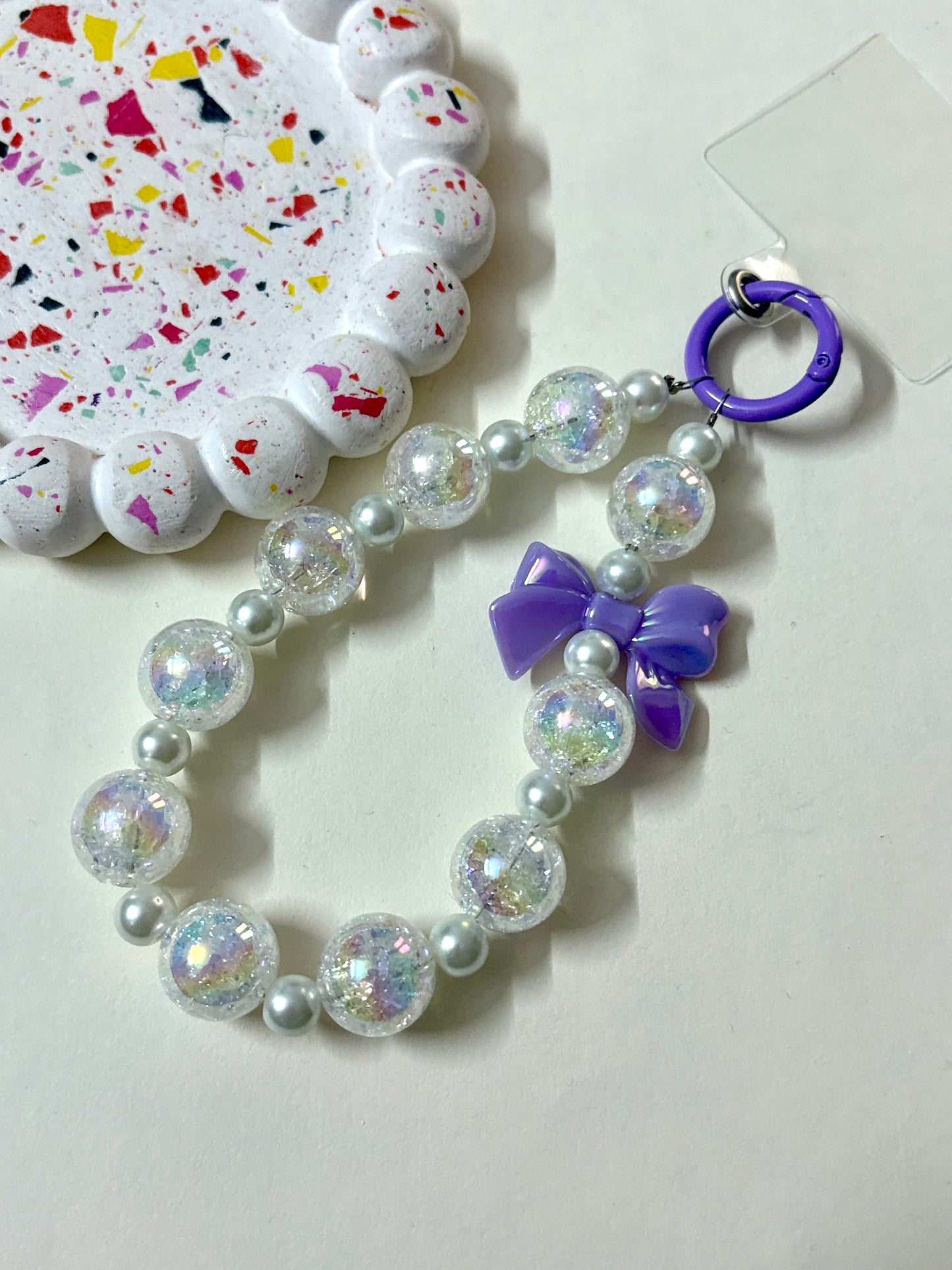 A stylish phone charm wrist strap featuring iridescent crackle beads, soft pearl accents, and a glossy black bow, attached to a black phone case.