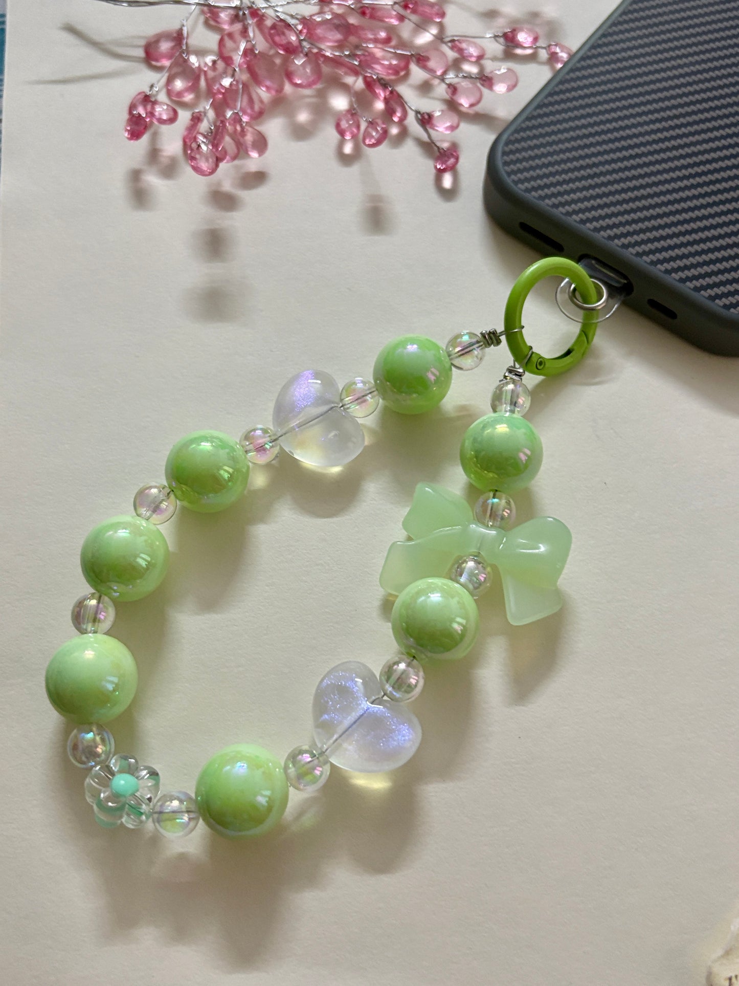 A cute phone charm made with glossy and translucent beads in various shapes, including a bow accent, strung together on an elastic loop with a plastic strap attachment.