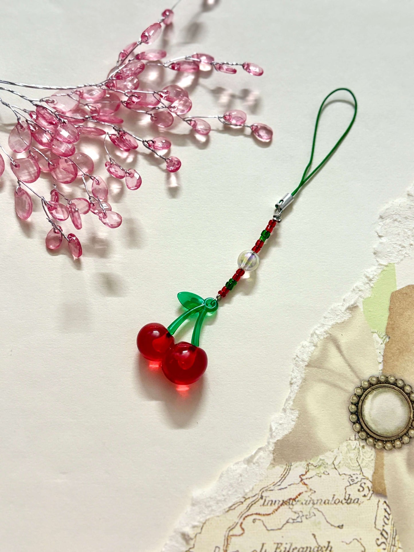 Playful phone charm featuring a glossy cherry pendant with green stem detail, connected by a beaded strap and a loop for phone attachment.