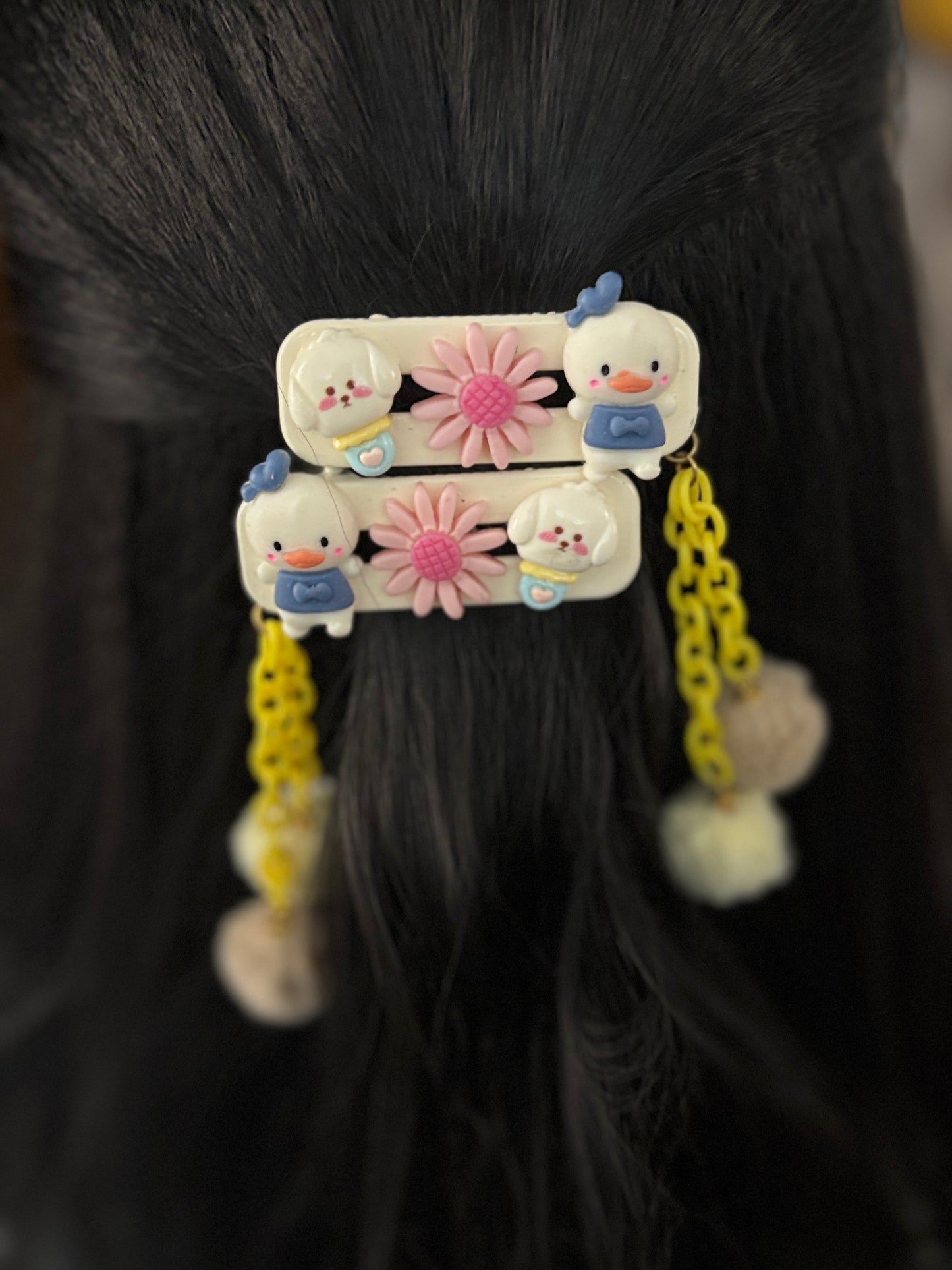 A pair of cute hair clips featuring Donald Duck, pink flowers, yellow plastic chains, and fuzzy pom pom dangles—fun and playful cartoon hair accessory.
