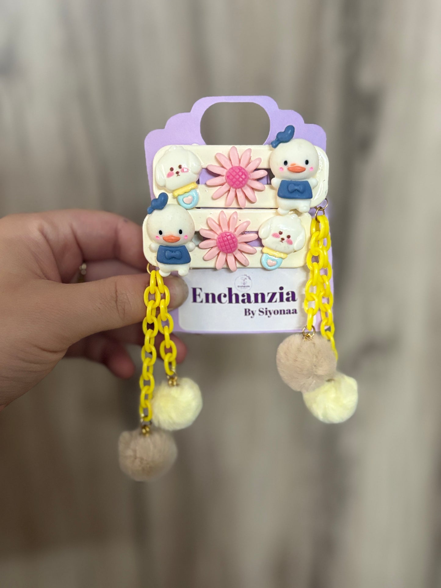 A pair of cute hair clips featuring Donald Duck, pink flowers, yellow plastic chains, and fuzzy pom pom dangles—fun and playful cartoon hair accessory.