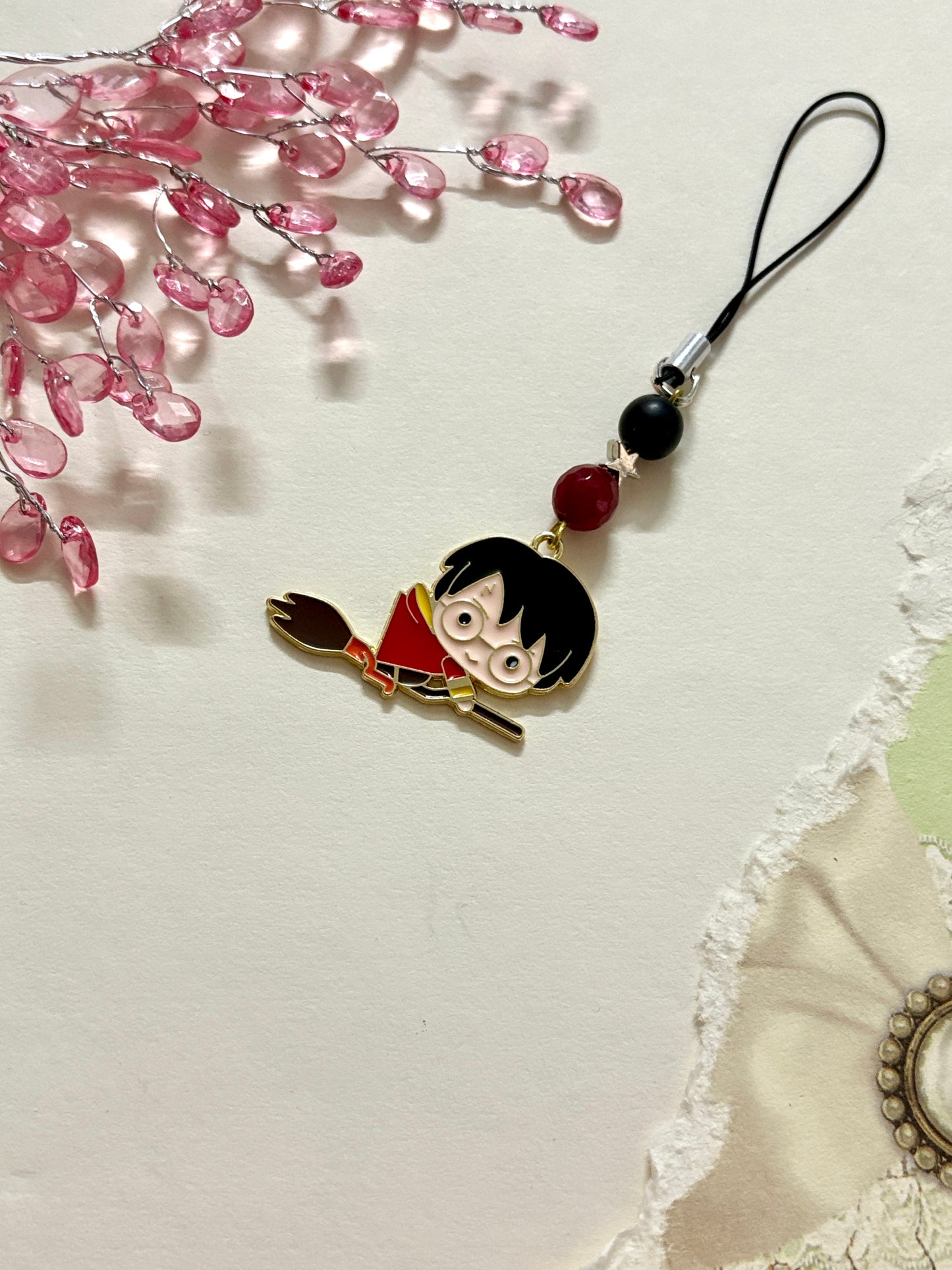 A Harry Potter phone charm featuring a cartoon-style Harry flying on a broomstick, adorned with black and maroon beads and a translucent bow on a black strap.