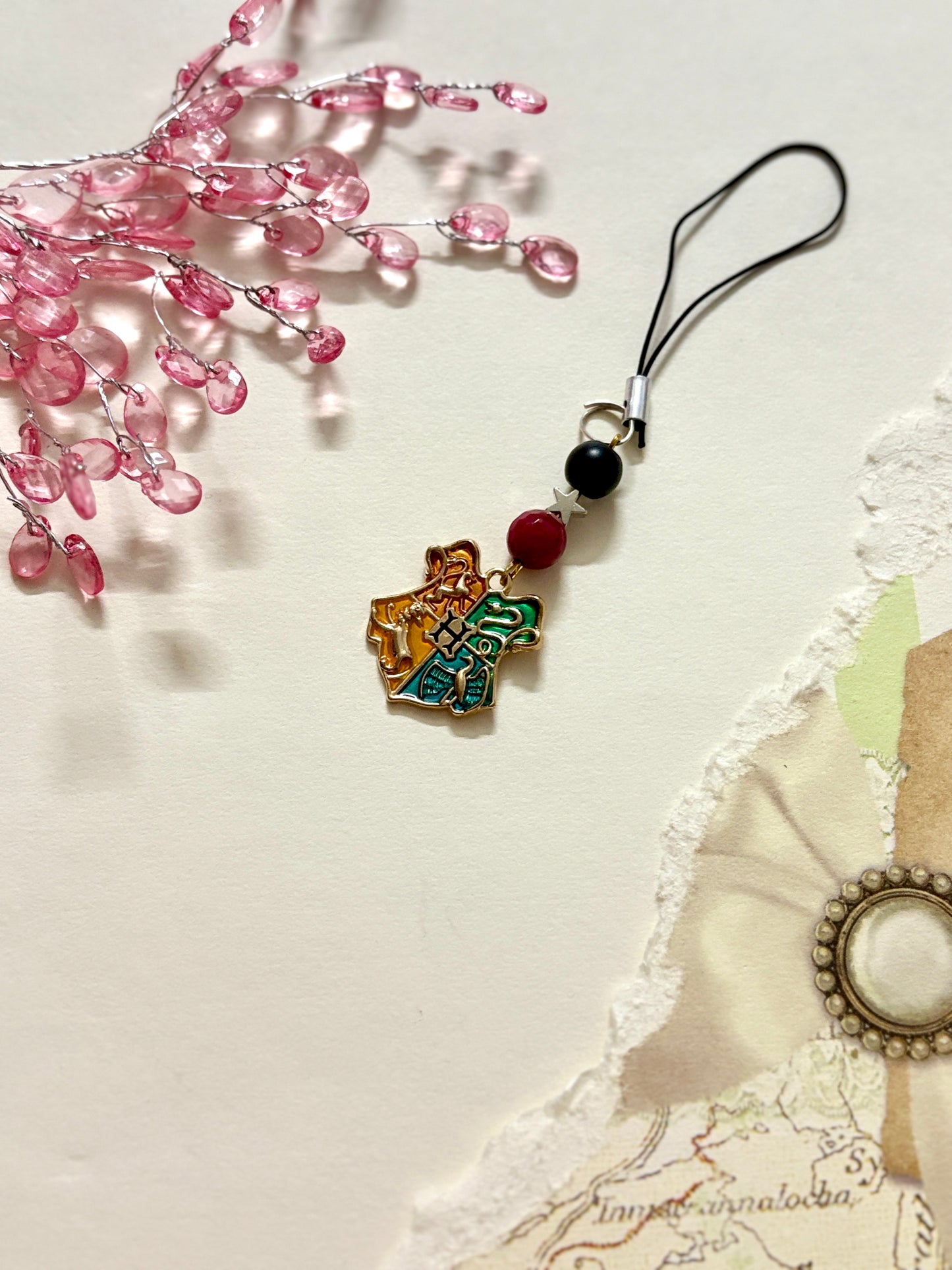 A Hogwarts crest-themed phone charm with a gold enamel charm showcasing the four house symbols, paired with a clear bow and black strap.