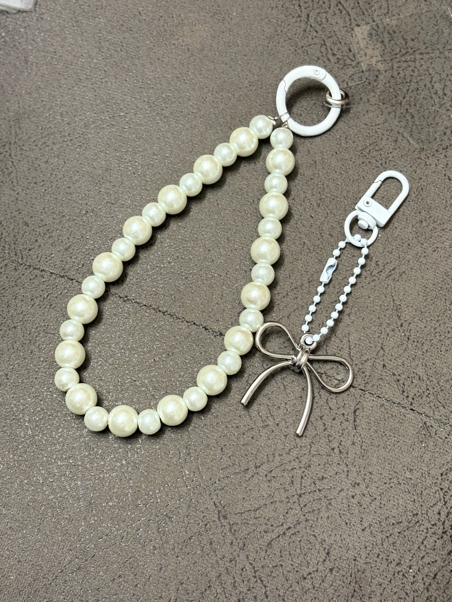 Elegant phone charm made with glossy pearl-like beads and a minimalist metallic bow charm, displayed on a textured background.