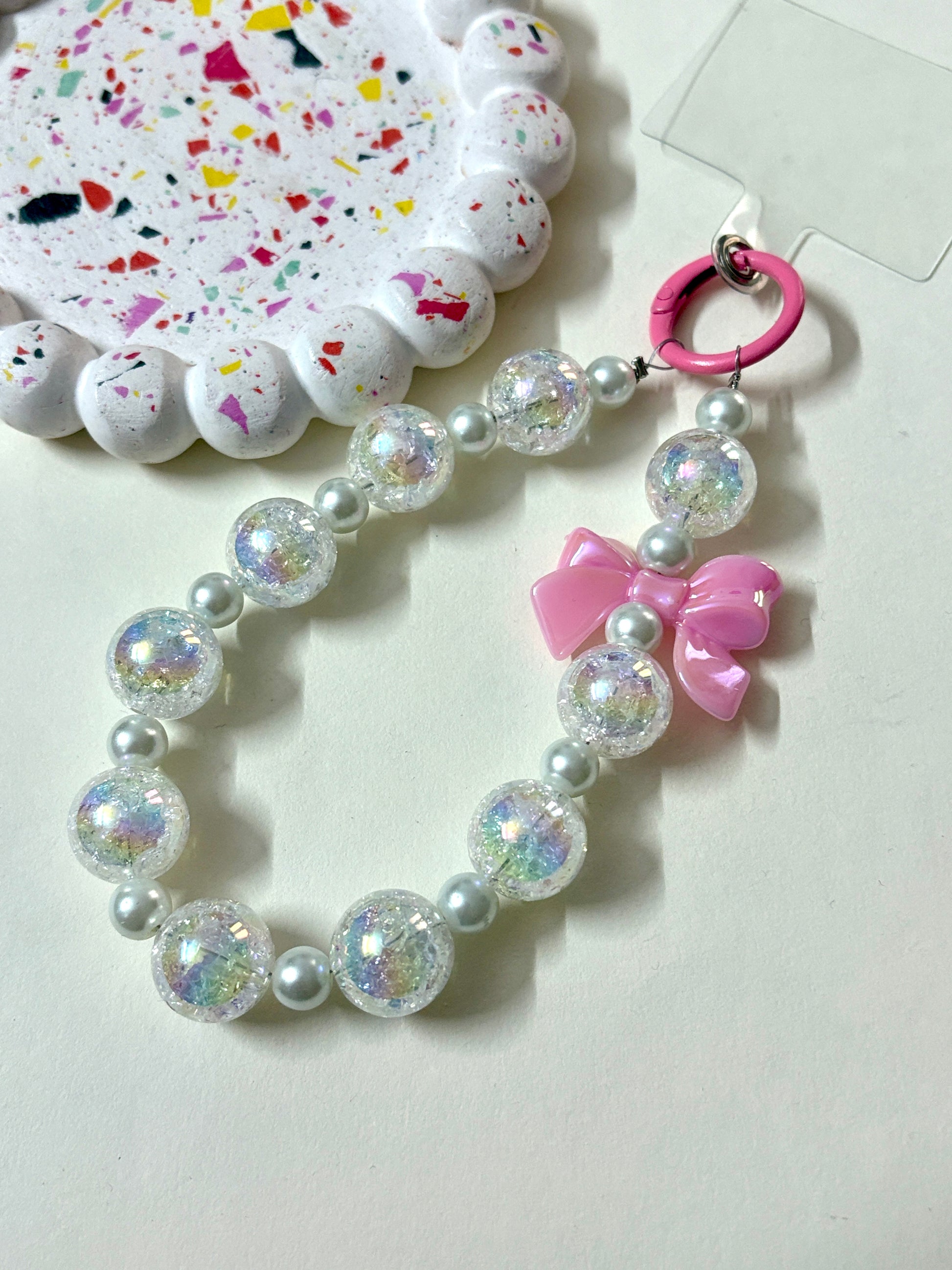A stylish phone charm wrist strap featuring iridescent crackle beads, soft pearl accents, and a glossy black bow, attached to a black phone case.