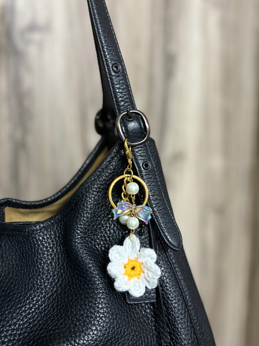 Bag charm featuring a crochet flower with pearl beads and a bow, attached to a black leather bag. Ideal for adding a soft, handmade touch to accessories.