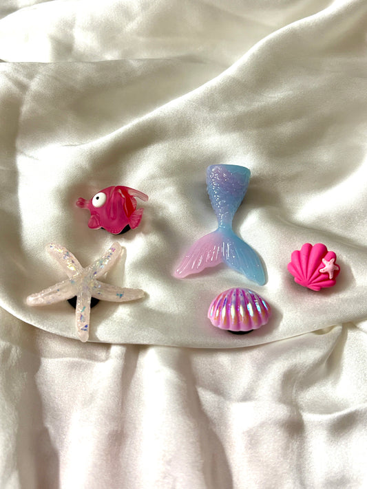 Set of 5 ocean-themed Crocs Jibbitz charms including mermaid tail, glittery starfish, pink fish, and vibrant shell designs—perfect for beachy vibes.