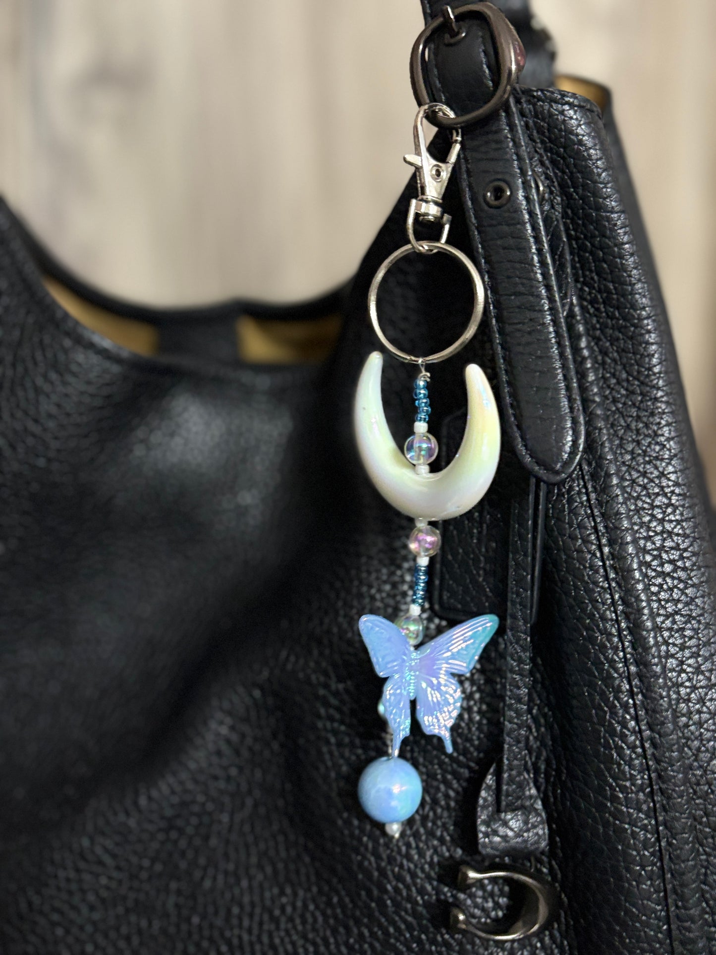Elegant bag charm with moon, butterfly, and bead details hanging from a black leather handbag. Adds a whimsical and stylish touch to any bag.