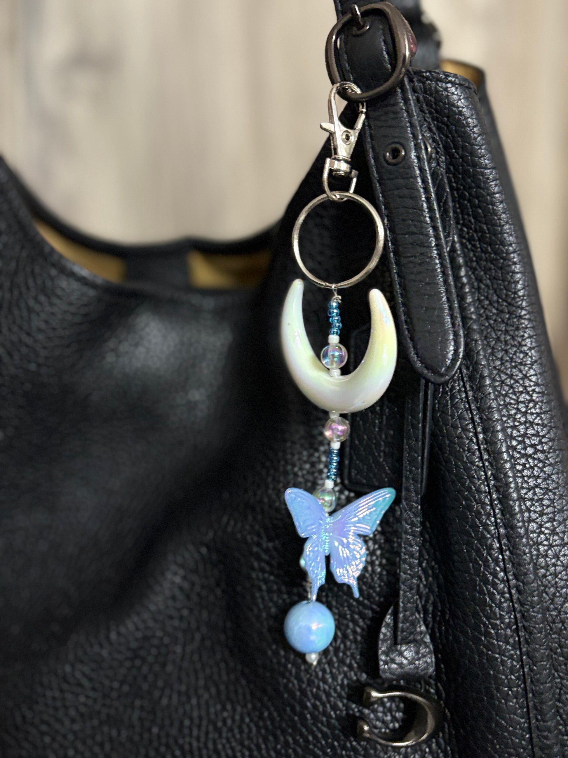 Elegant bag charm with moon, butterfly, and bead details hanging from a black leather handbag. Adds a whimsical and stylish touch to any bag.