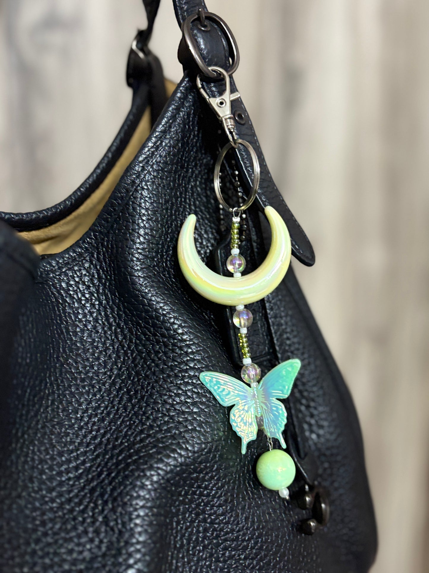Elegant bag charm with moon, butterfly, and bead details hanging from a black leather handbag. Adds a whimsical and stylish touch to any bag.