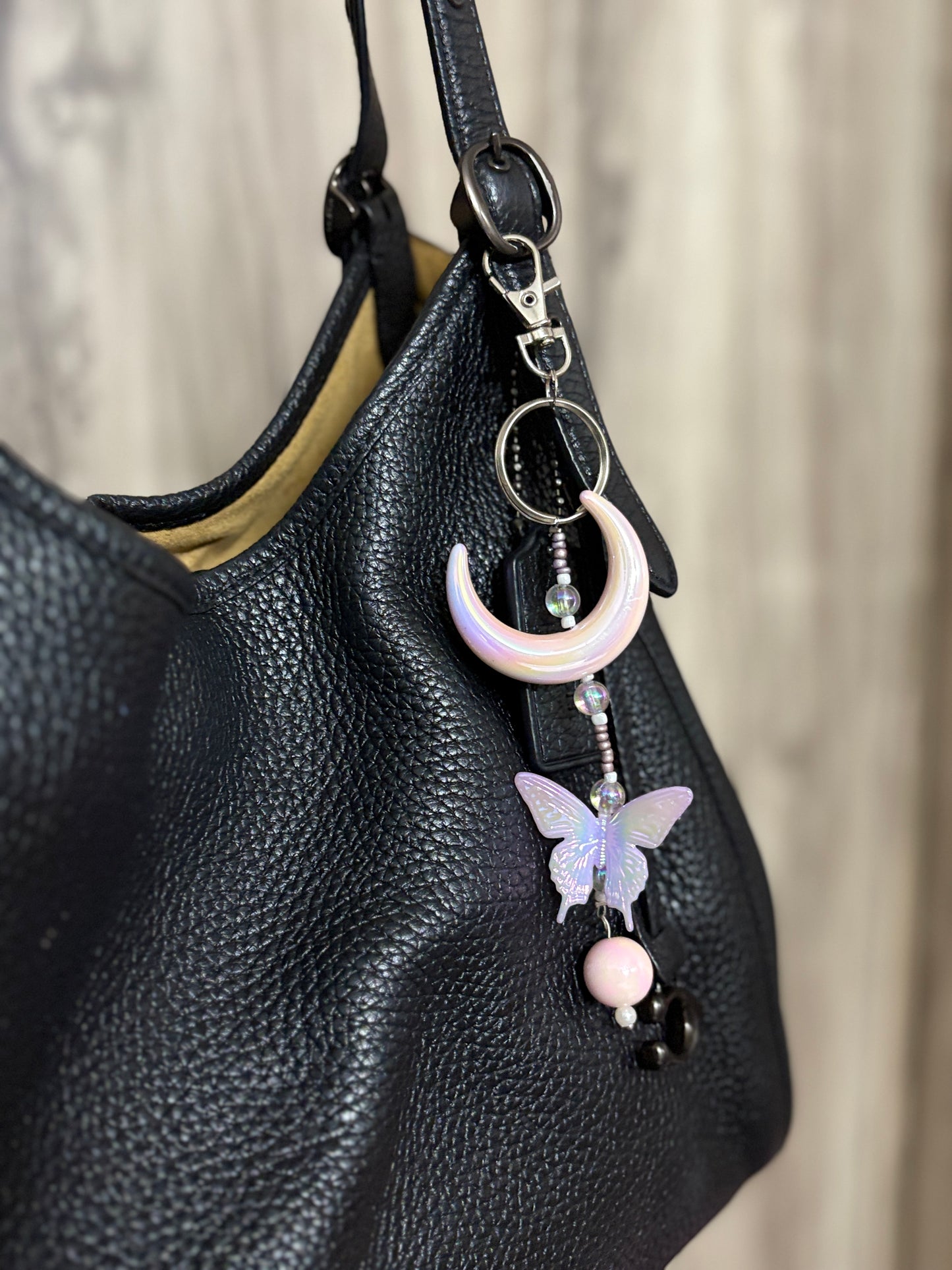 Elegant bag charm with moon, butterfly, and bead details hanging from a black leather handbag. Adds a whimsical and stylish touch to any bag.