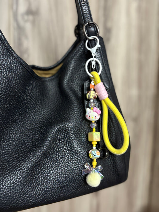 Bag charm featuring playful beads, cartoon charms, and a soft pompom with a loop attachment, hanging from a black leather handbag. Ideal for quirky styling.