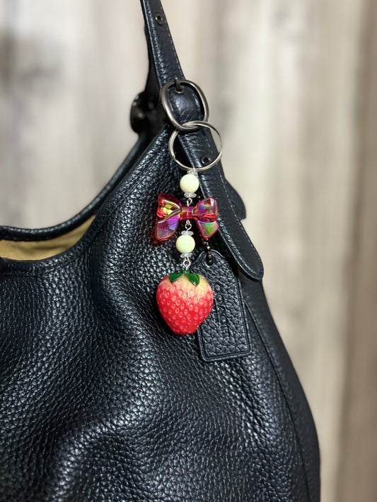 Cute bag charm with a strawberry pendant, bow, and beads hanging from a black leather handbag. Ideal for accessorizing bags with a fun, playful touch.