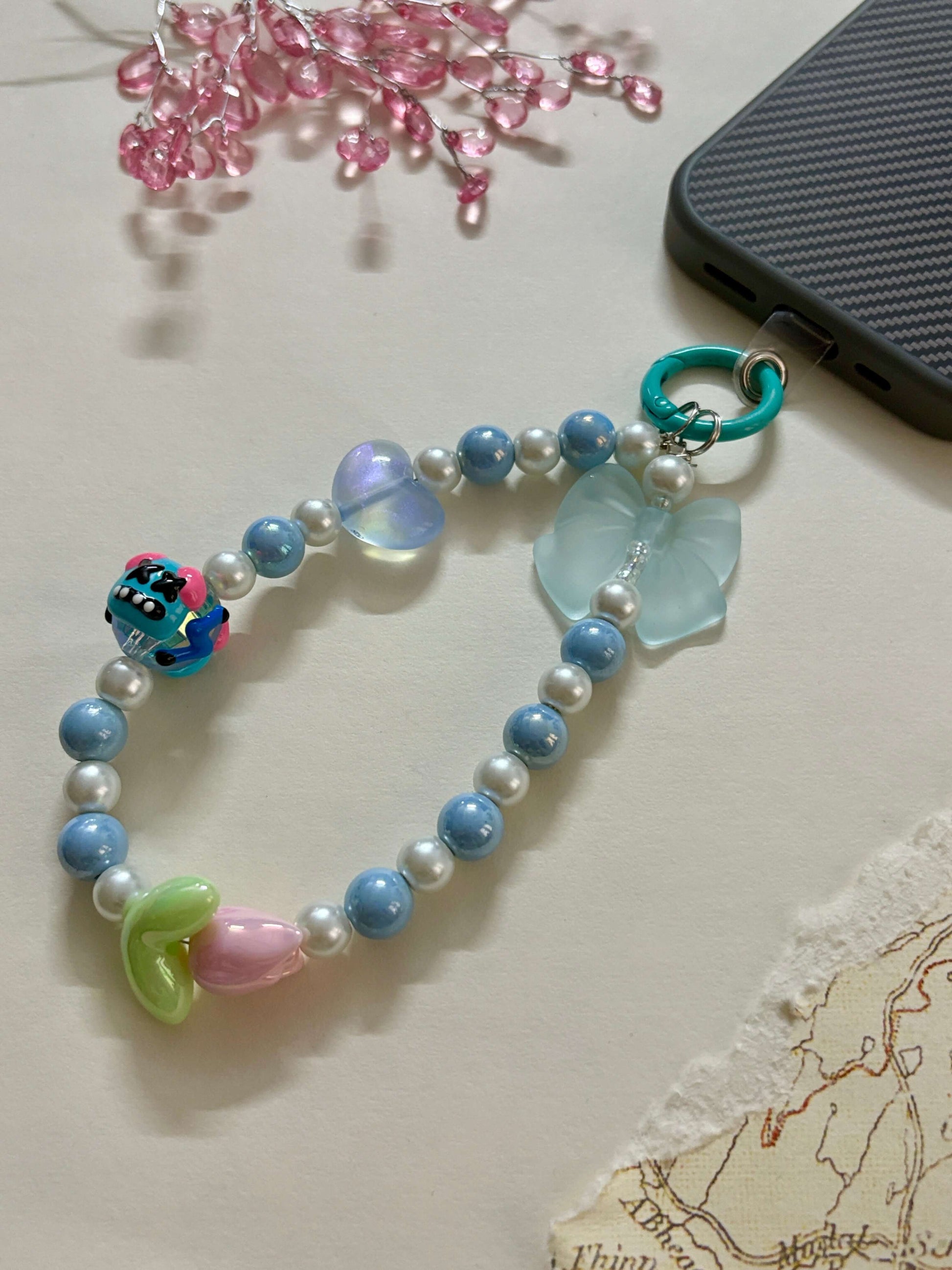 A beaded phone charm strap featuring glossy pearl-like beads, miniature character and flower charms, and a loop attachment; designed as a cute and functional phone accessory for personalization and added grip.