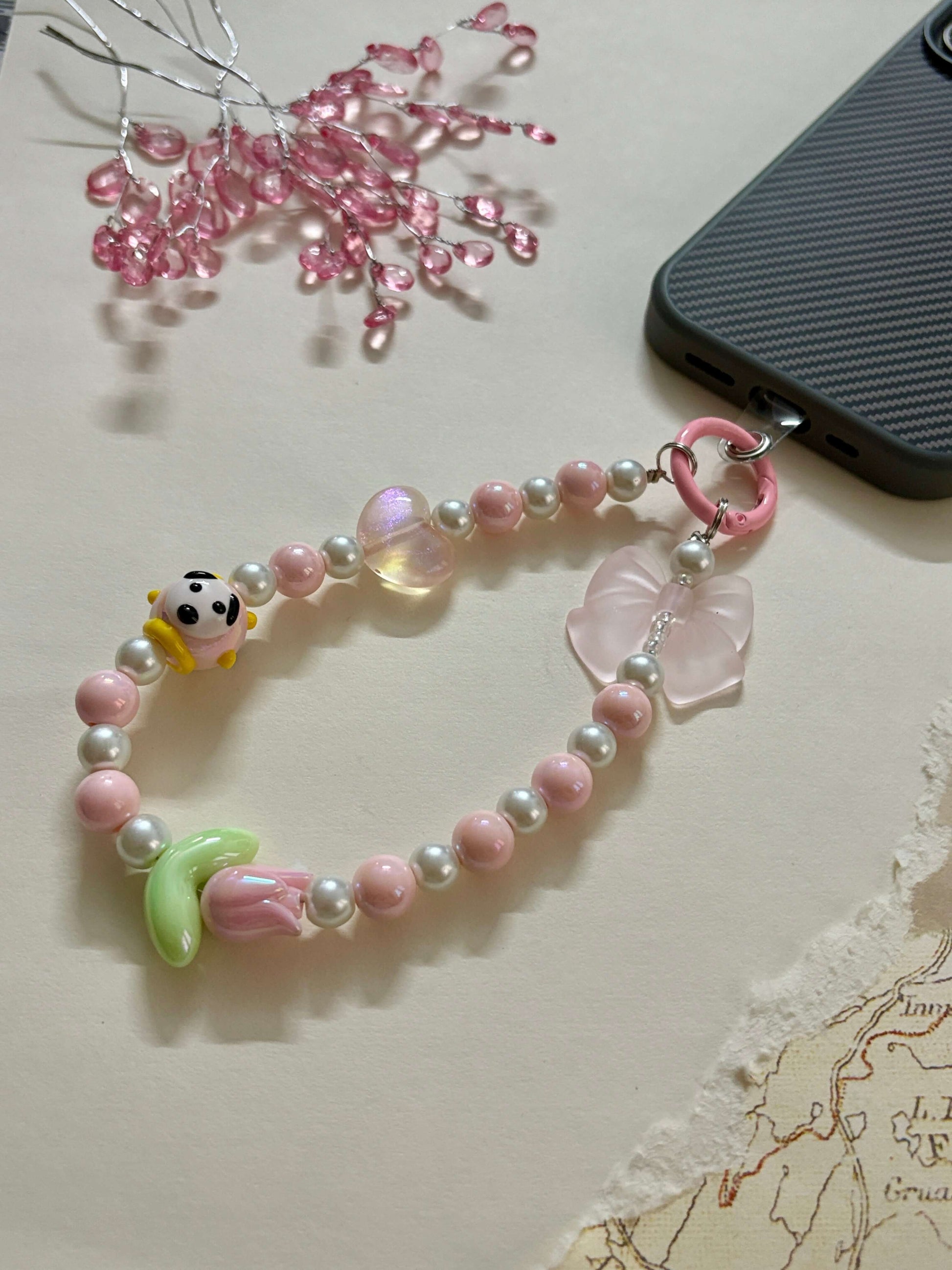 A beaded phone charm strap featuring glossy pearl-like beads, miniature character and flower charms, and a loop attachment; designed as a cute and functional phone accessory for personalization and added grip.