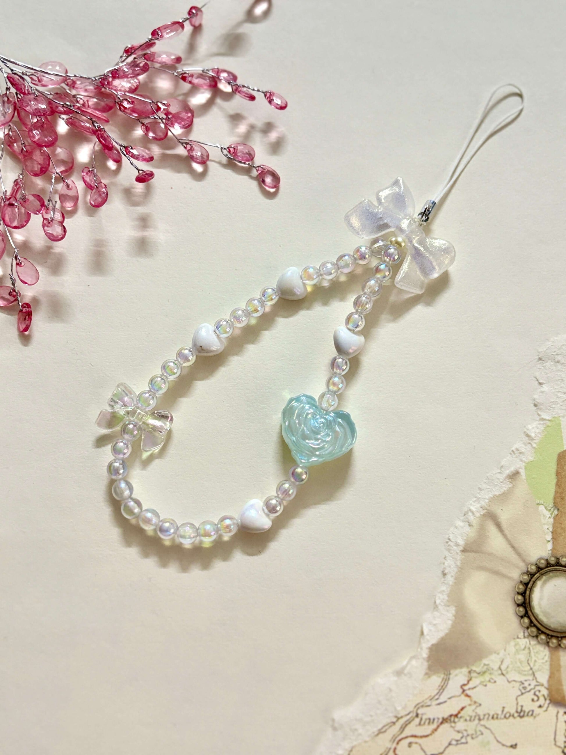 Beaded phone charm featuring a rose-shaped heart, delicate bow, and small heart accents; designed for attaching to mobile phones, adding a stylish and cute touch.