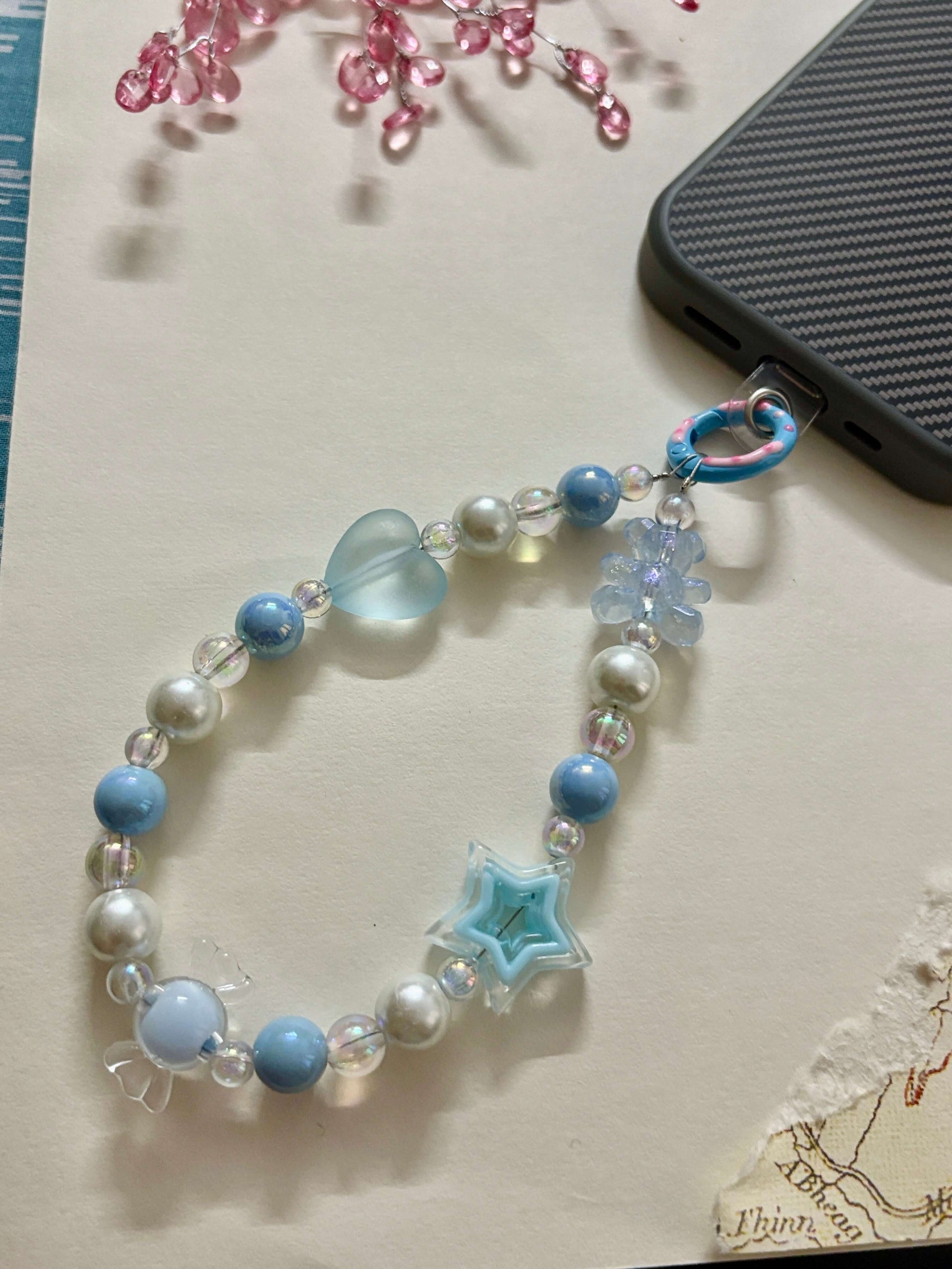Beaded phone charm with heart, star, and teddy bear accents, designed to hang from a mobile device; features pearl and translucent beads for a cute and playful look.