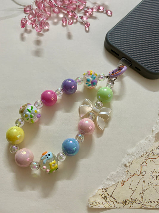 Colorful pastel beaded phone charm wrist strap featuring kawaii bunny and cupcake beads, clear accent beads, and a glossy white bow, attached to a phone case.