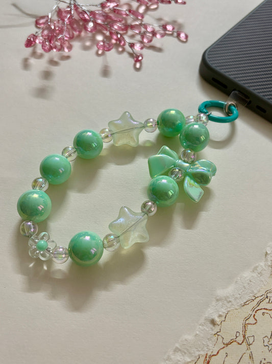 A cute phone charm made with glossy and translucent beads in various shapes, including a bow accent, strung together on an elastic loop with a plastic strap attachment.
