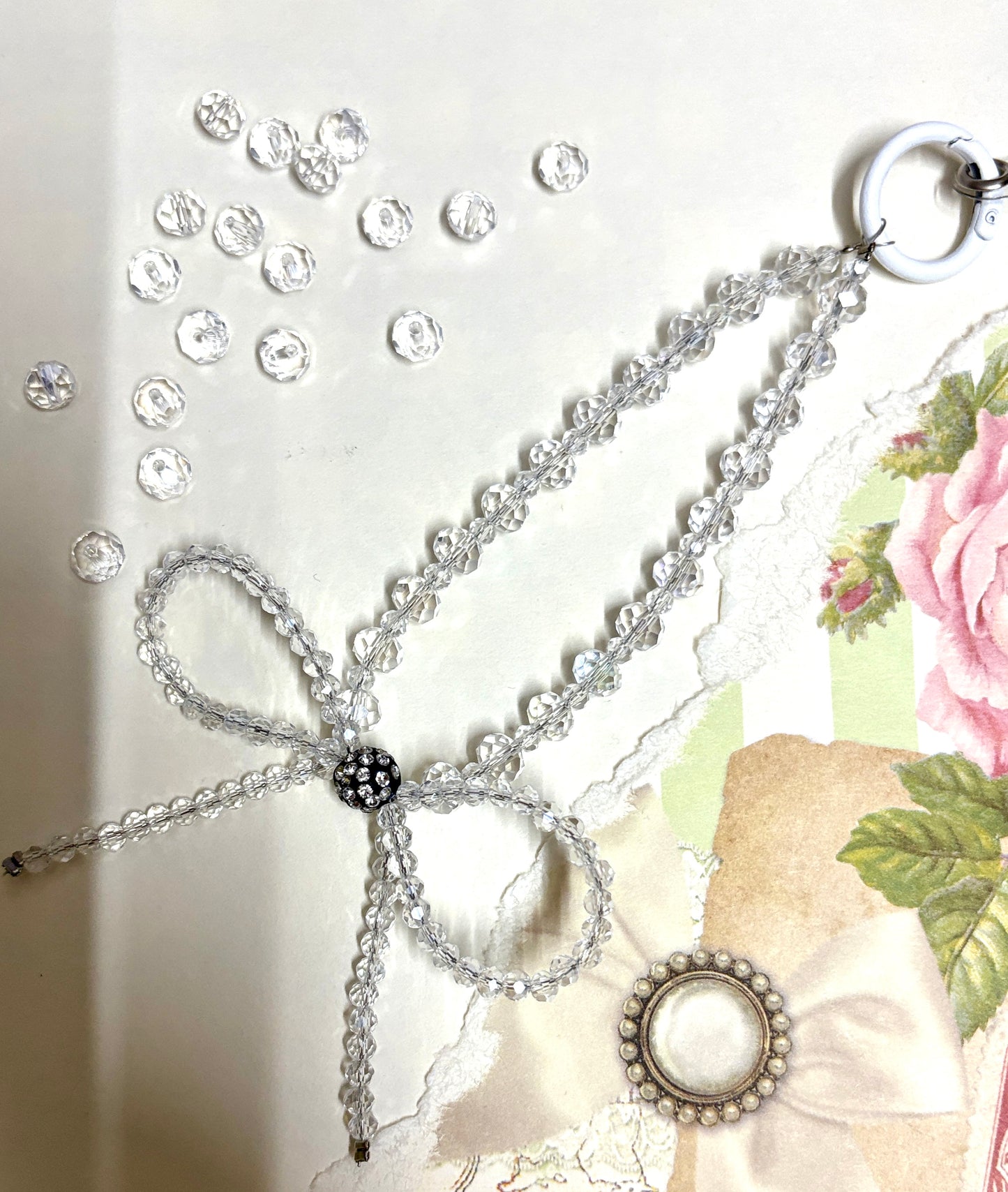 Elegant beaded phone charm featuring a delicate crystal bow design with faceted beads and a rhinestone center, styled with vintage floral paper.