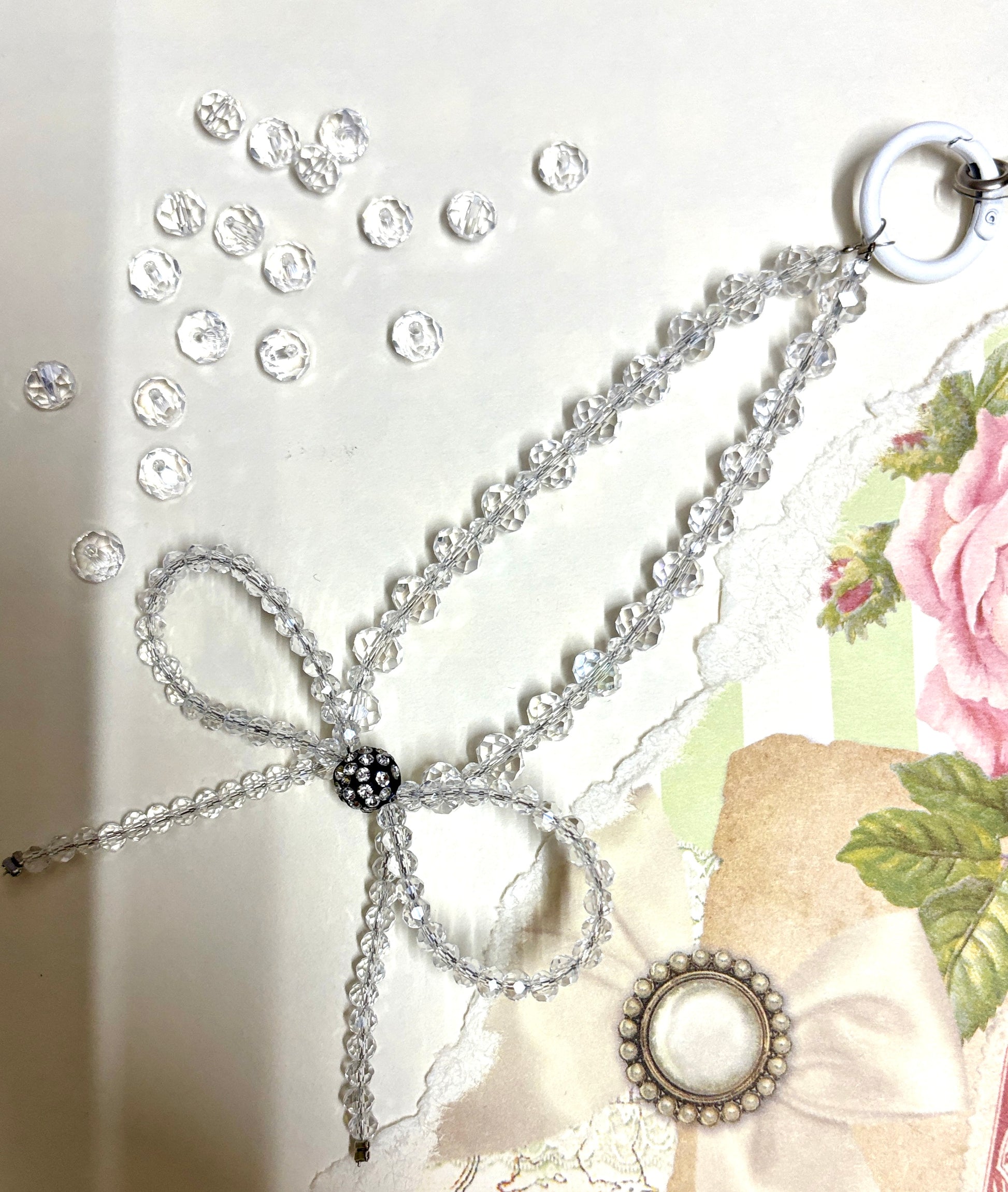 Elegant beaded phone charm featuring a delicate crystal bow design with faceted beads and a rhinestone center, styled with vintage floral paper.