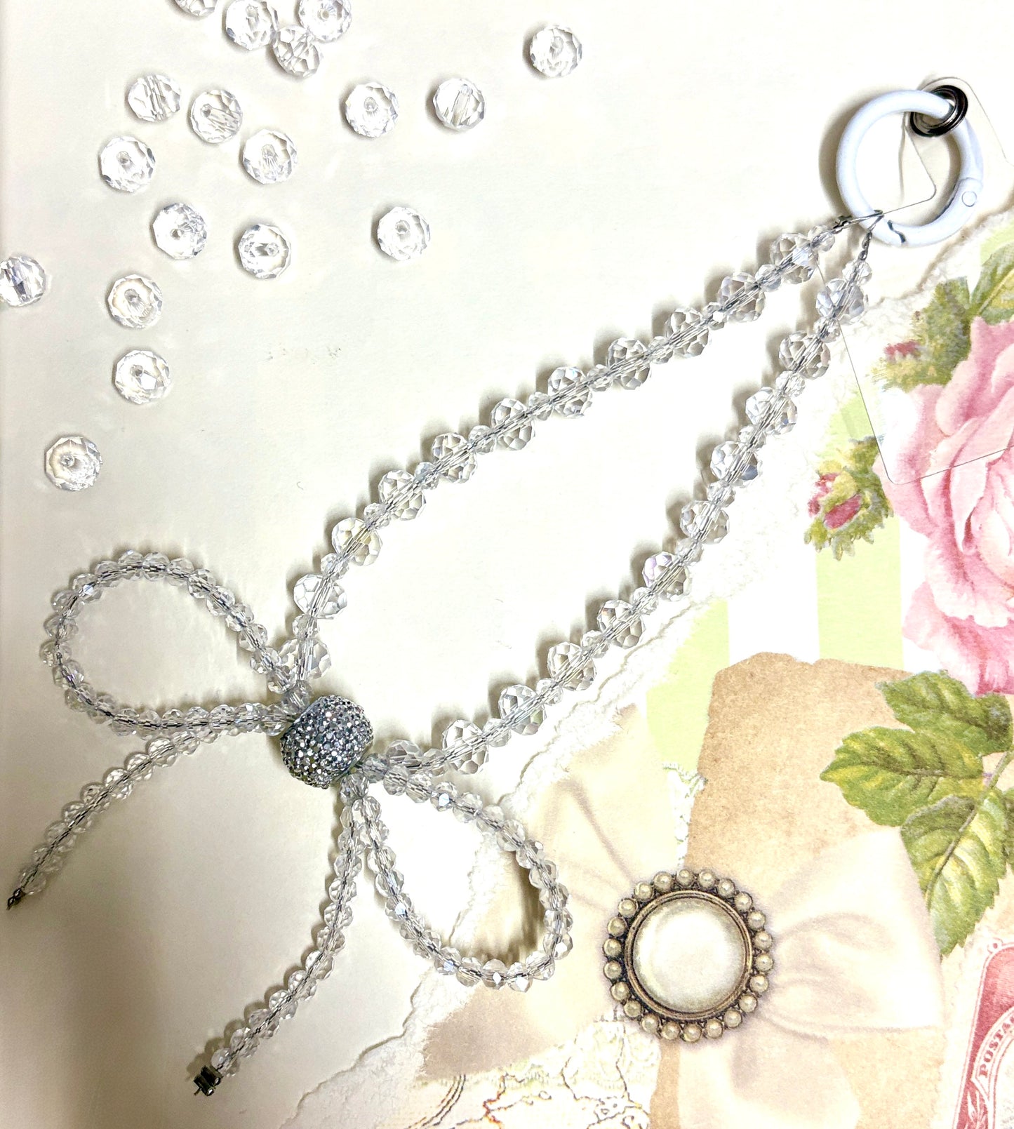 Elegant beaded phone charm featuring a delicate crystal bow design with faceted beads and a rhinestone center, styled with vintage floral paper.