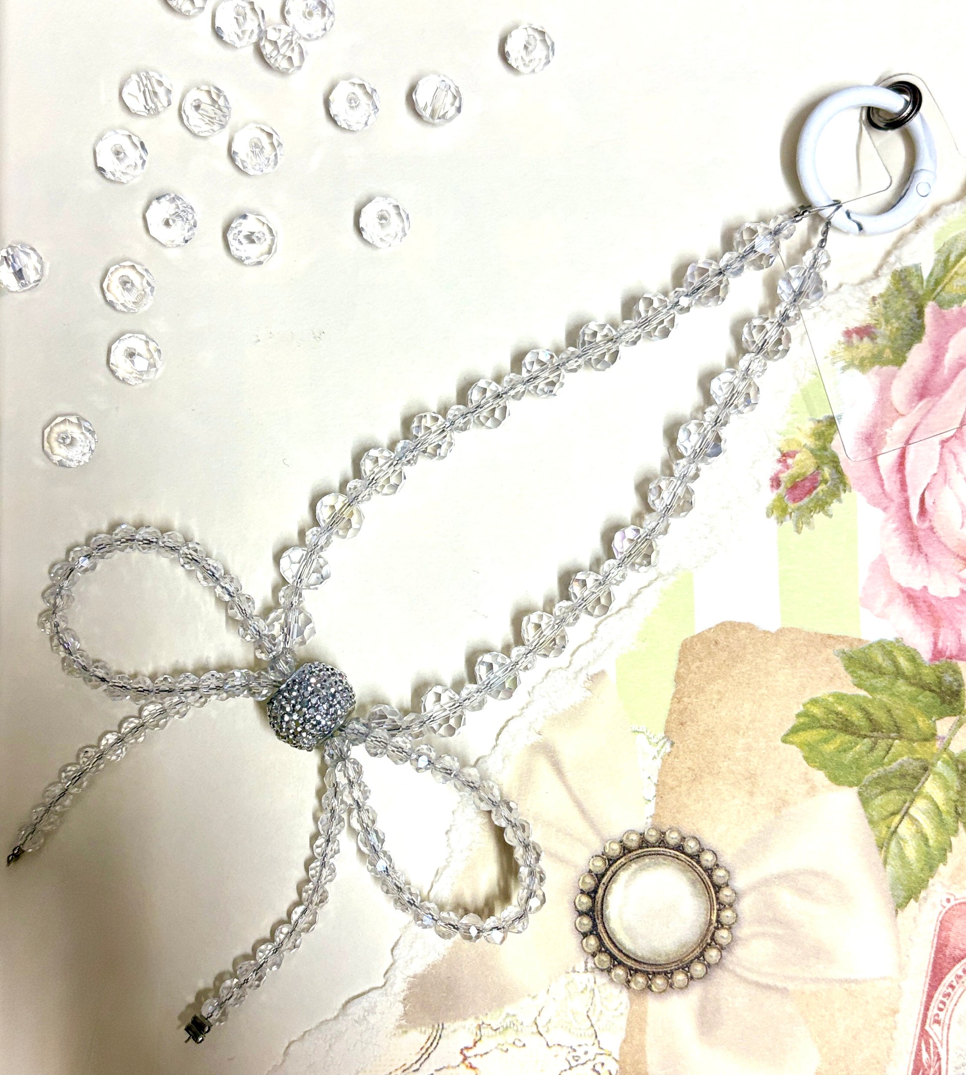 Elegant beaded phone charm featuring a delicate crystal bow design with faceted beads and a rhinestone center, styled with vintage floral paper.