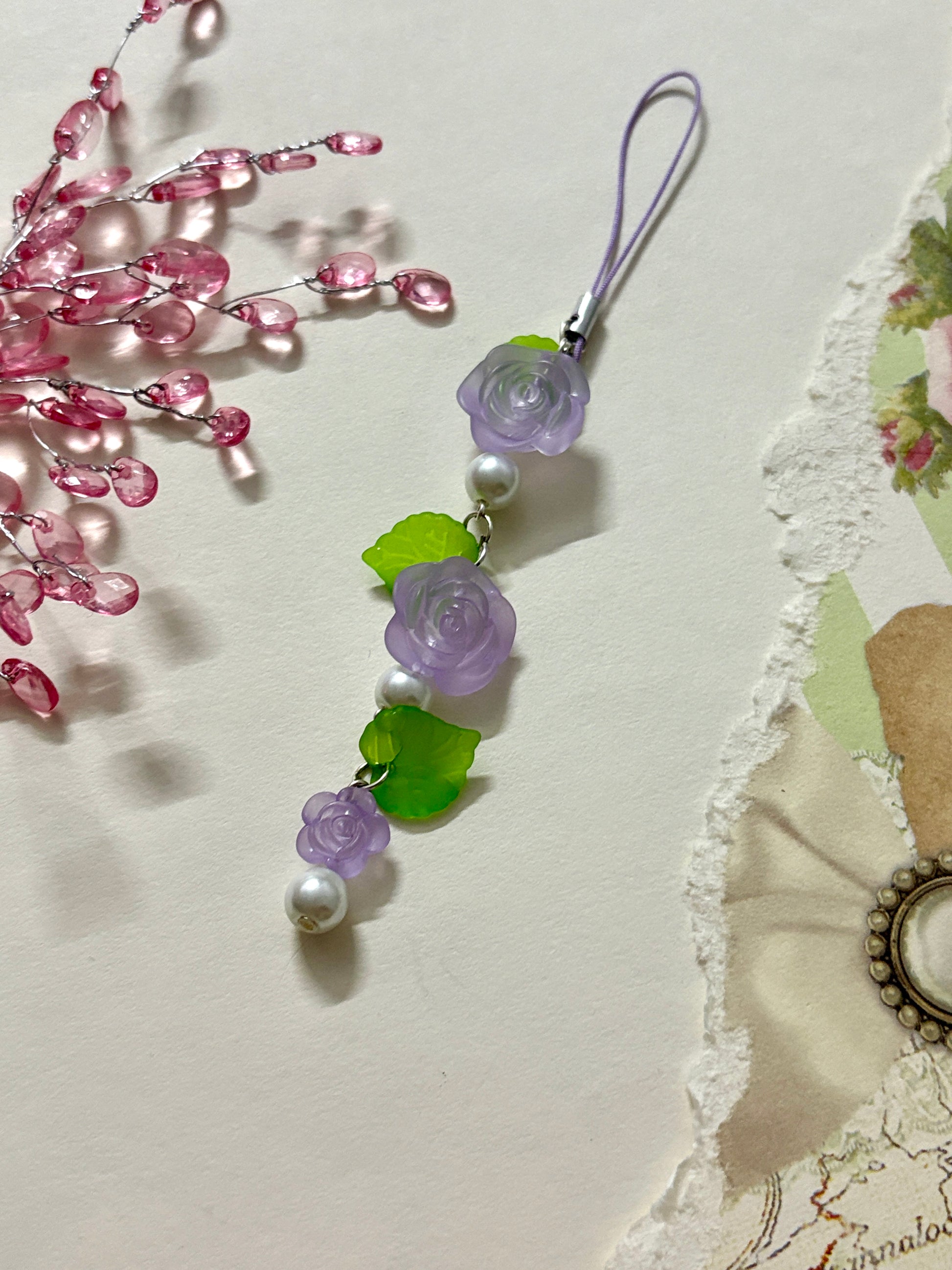 Floral phone charm featuring delicate resin roses, green leaves, and pearl accents. A graceful accessory available in four color variations.