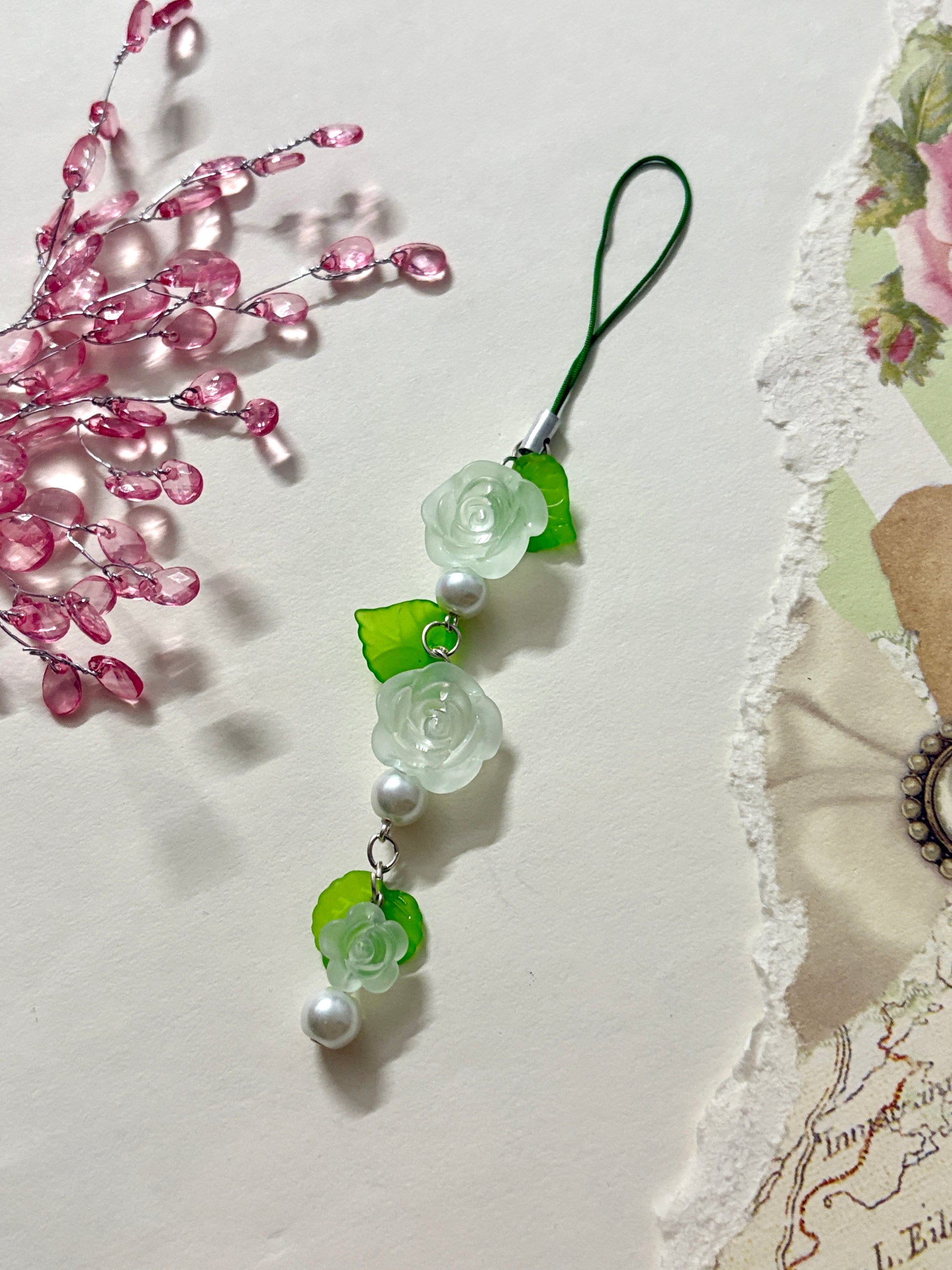Floral phone charm featuring delicate resin roses, green leaves, and pearl accents. A graceful accessory available in four color variations.