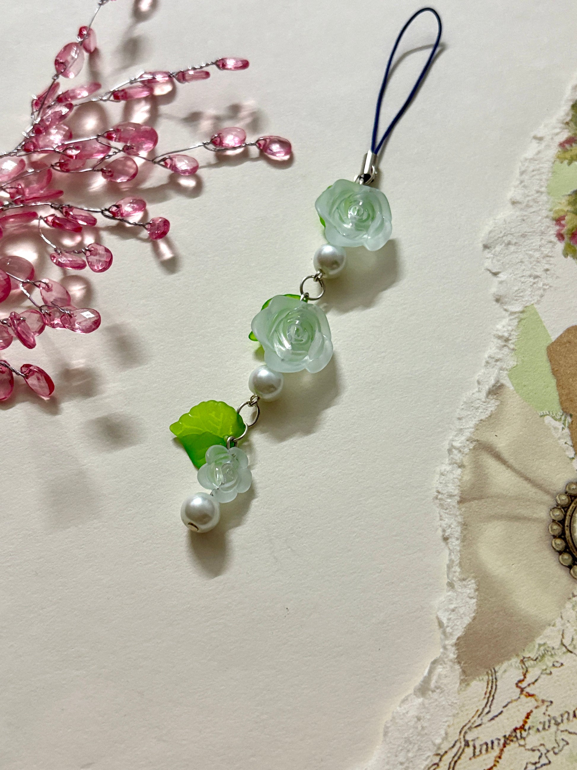 Floral phone charm featuring delicate resin roses, green leaves, and pearl accents. A graceful accessory available in four color variations.