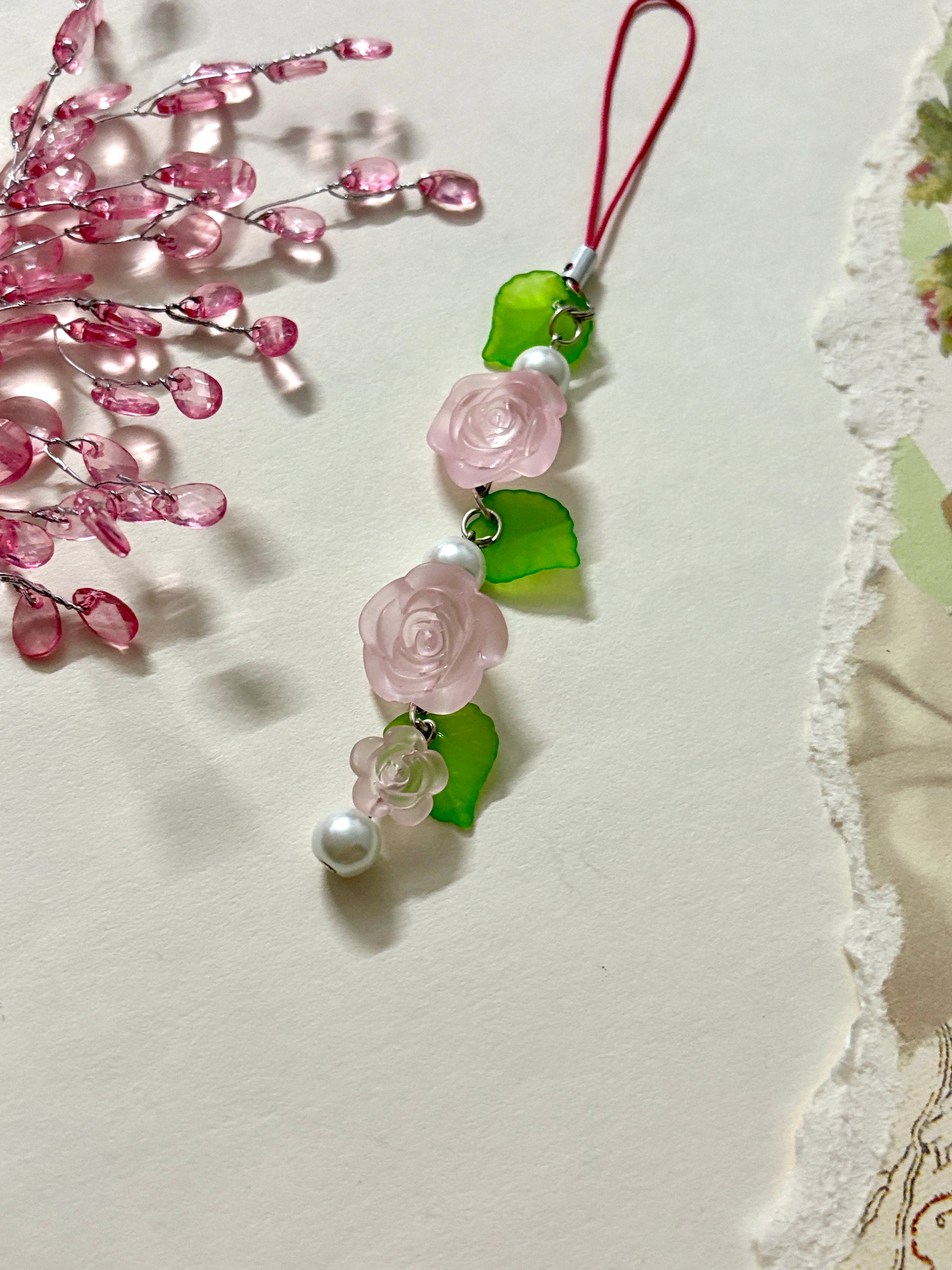 Floral phone charm featuring delicate resin roses, green leaves, and pearl accents. A graceful accessory available in four color variations.