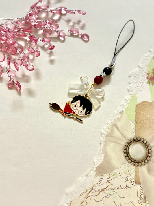 A Harry Potter phone charm featuring a cartoon-style Harry flying on a broomstick, adorned with black and maroon beads and a translucent bow on a black strap.