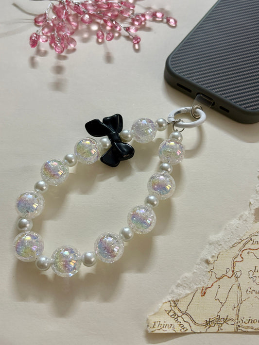 A stylish phone charm wrist strap featuring iridescent crackle beads, soft pearl accents, and a glossy black bow, attached to a black phone case.