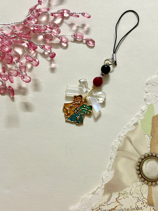 A Hogwarts crest-themed phone charm with a gold enamel charm showcasing the four house symbols, paired with a clear bow and black strap.