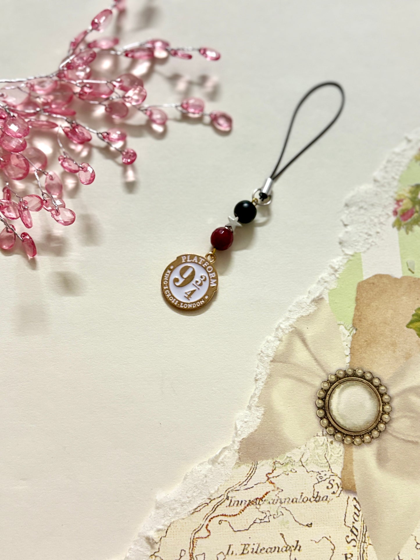 A phone charm inspired by Platform 9¾ from Harry Potter, featuring a white and gold enamel tag, clear bow, and beaded accents on a black strap.