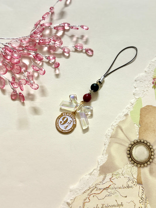 A phone charm inspired by Platform 9¾ from Harry Potter, featuring a white and gold enamel tag, clear bow, and beaded accents on a black strap.