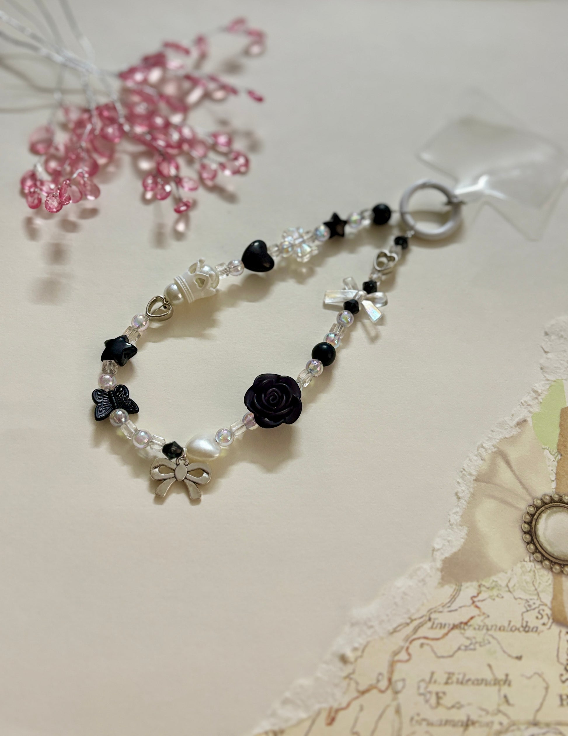 
“Handmade black and white beaded phone charm with rose, heart, bow, and butterfly charms in kawaii aesthetic”