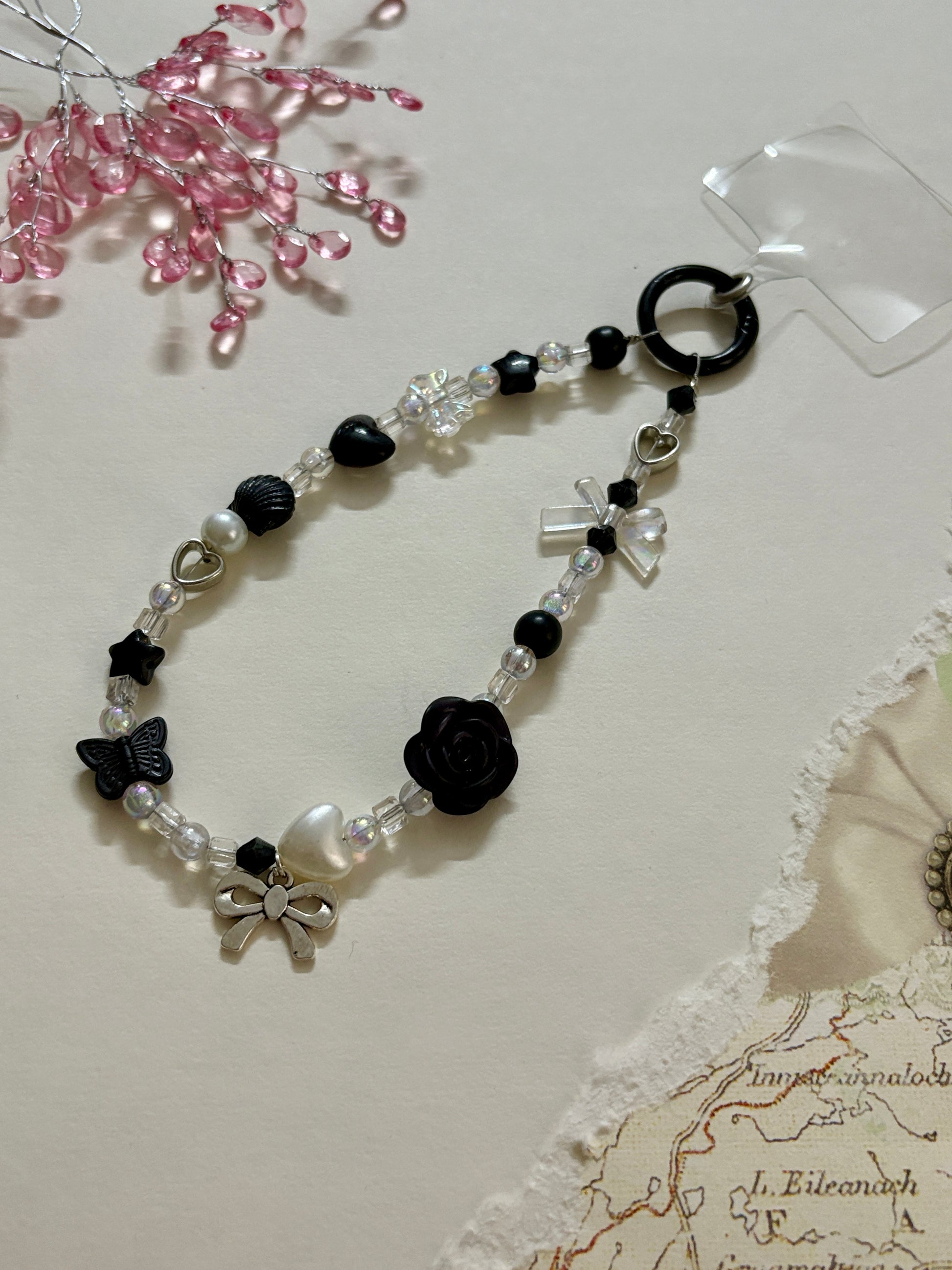 
“Handmade black and white beaded phone charm with rose, heart, bow, and butterfly charms in kawaii aesthetic”