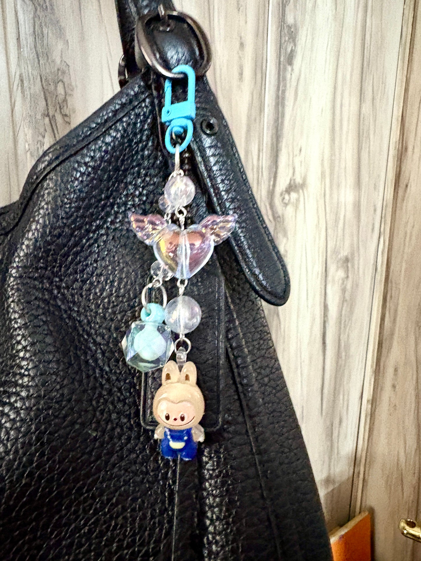 Labubu bag charm featuring a blue-outfitted figure, crystal beads, and pastel clip — a rare and playful accessory available in 2 colors, attached to a black bag.
