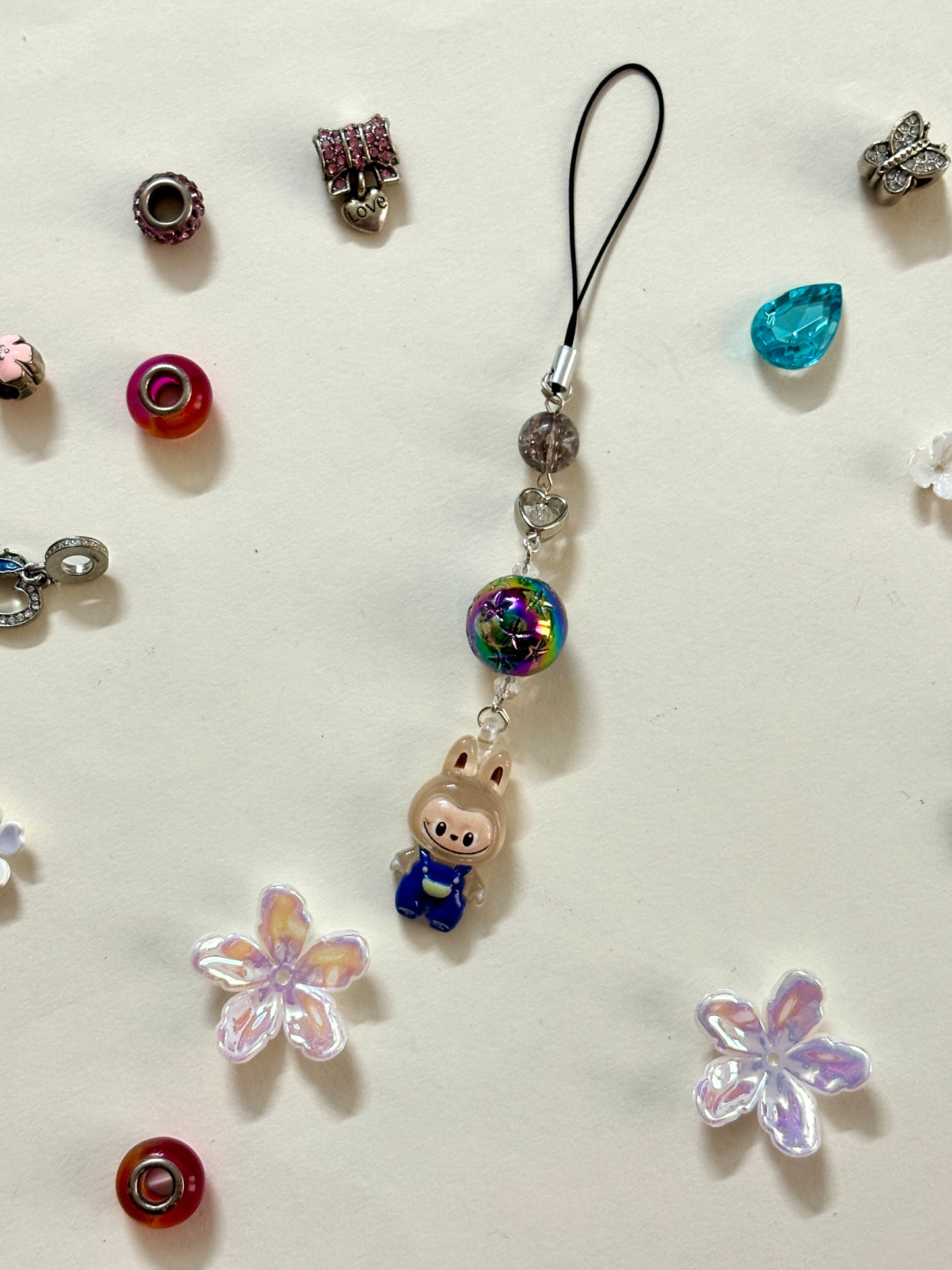 Mini Labubu phone charm with rainbow orb bead and black strap, featuring Labubu in blue overalls — collectible strap available in two color options.