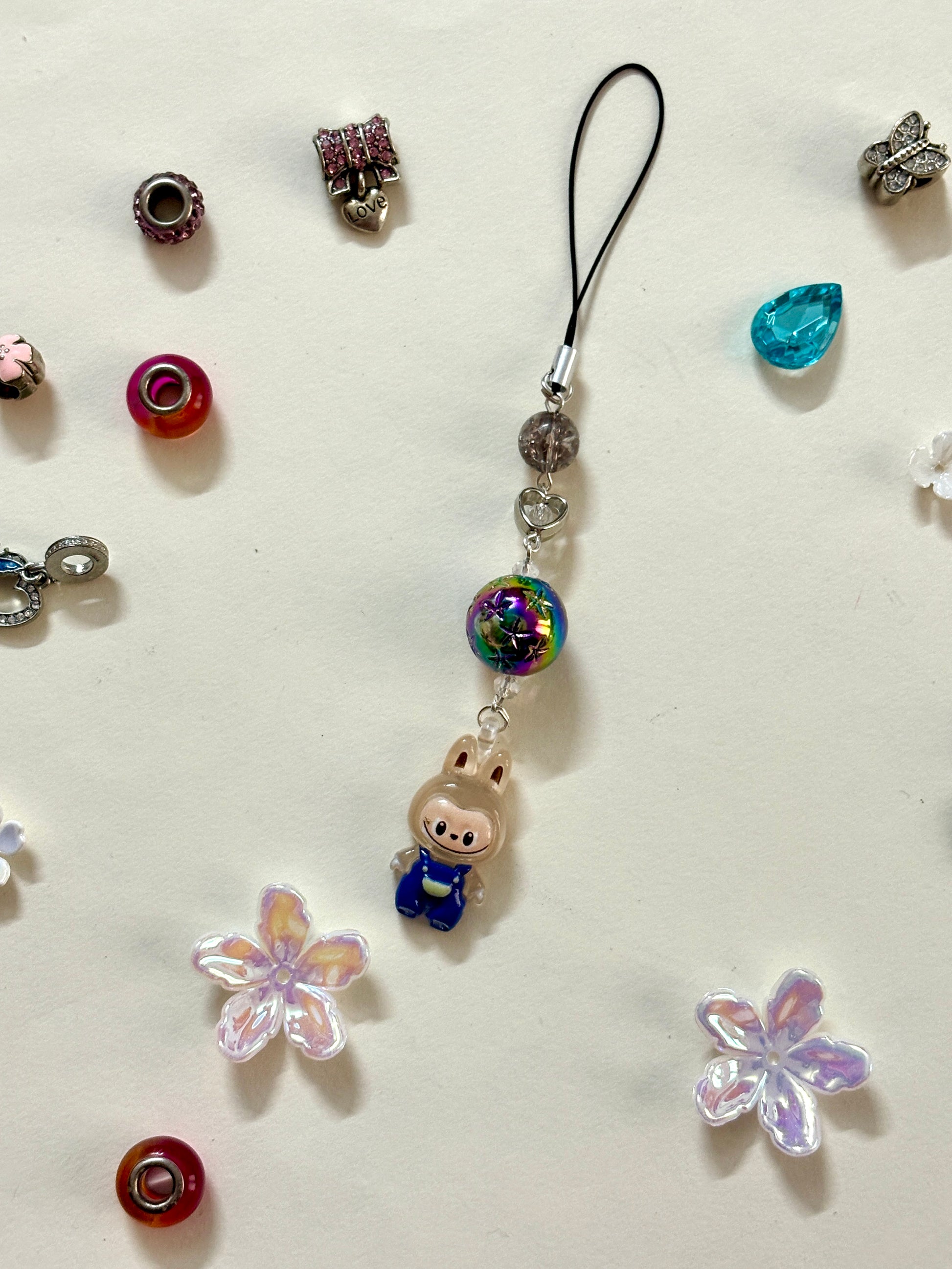 Mini Labubu phone charm with rainbow orb bead and black strap, featuring Labubu in blue overalls — collectible strap available in two color options.