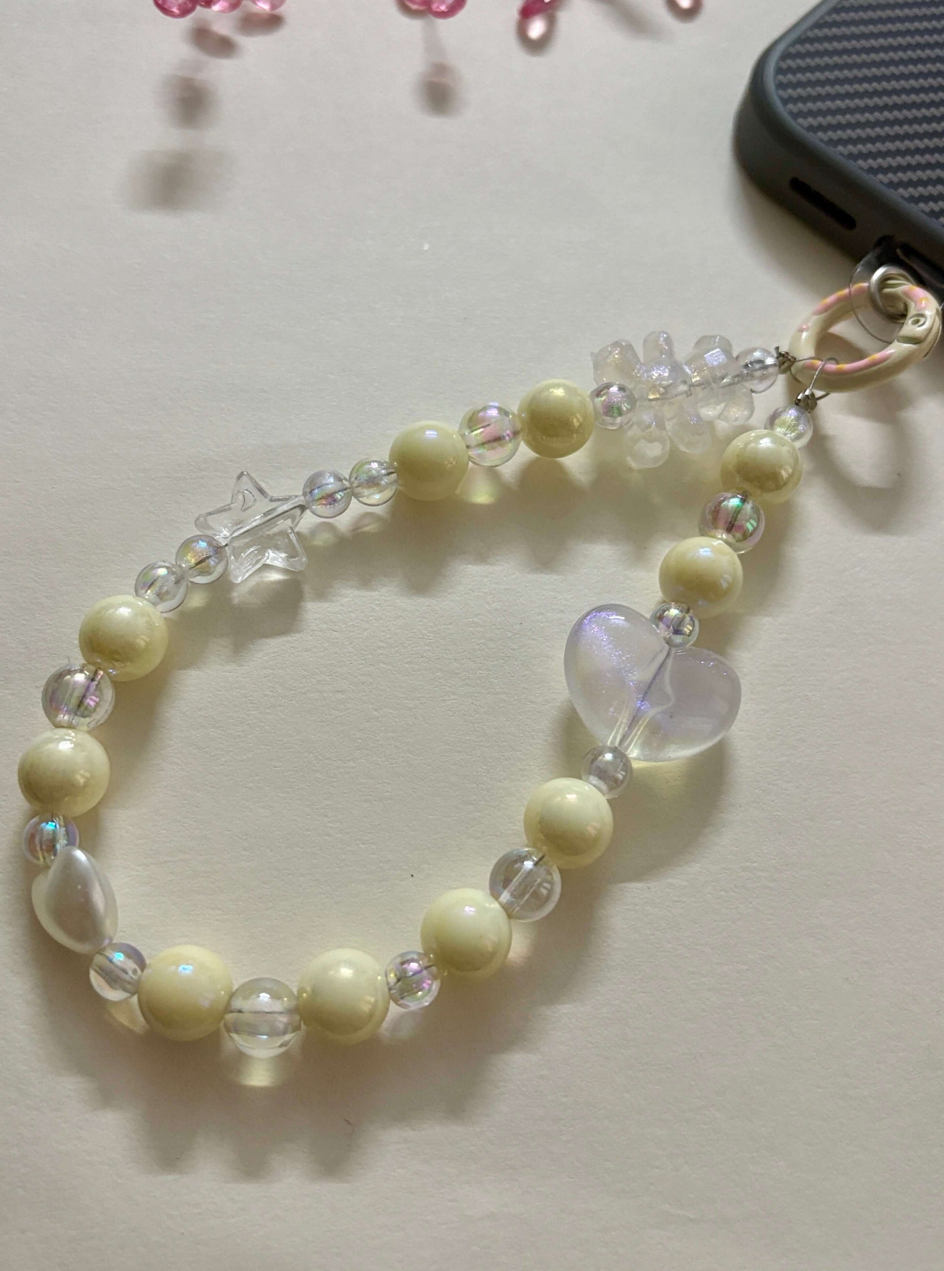 Beaded phone charm with heart, star, and teddy bear accents, designed to hang from a mobile device; features pearl and translucent beads for a cute and playful look.