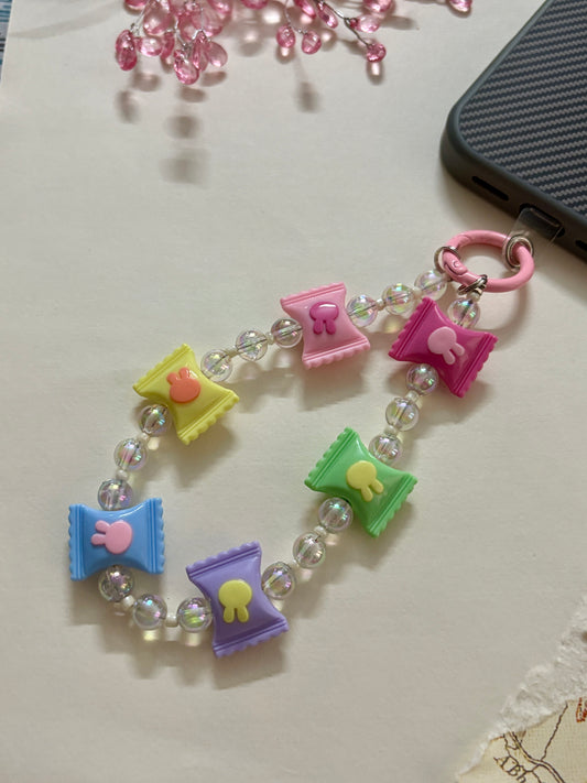 Cute pastel phone charm wrist strap featuring colorful candy-shaped beads in pink, purple, green, yellow, and blue with iridescent accent beads and a pink clasp attached to a phone case.
