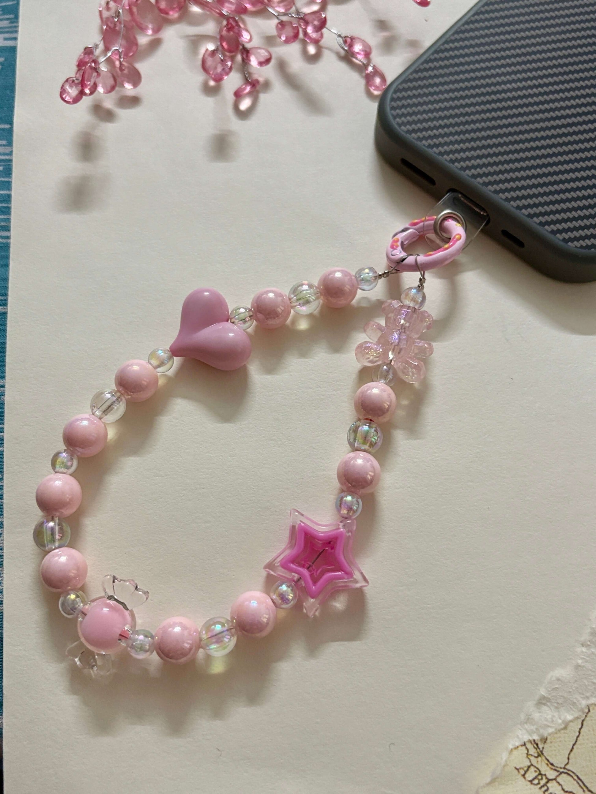 Beaded phone charm with heart, star, and teddy bear accents, designed to hang from a mobile device; features pearl and translucent beads for a cute and playful look.