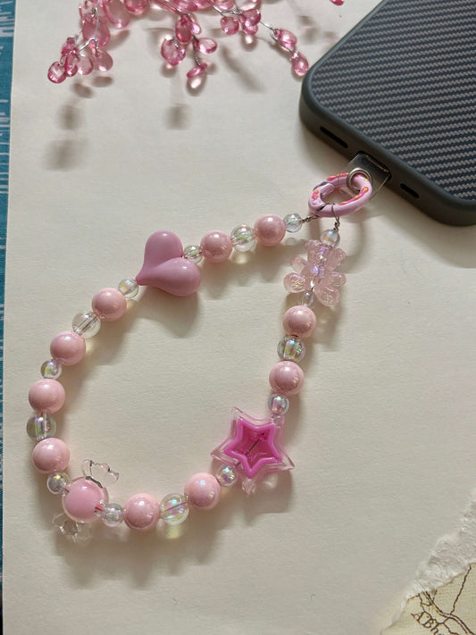 Beaded phone charm with heart, star, and teddy bear accents, designed to hang from a mobile device; features pearl and translucent beads for a cute and playful look.