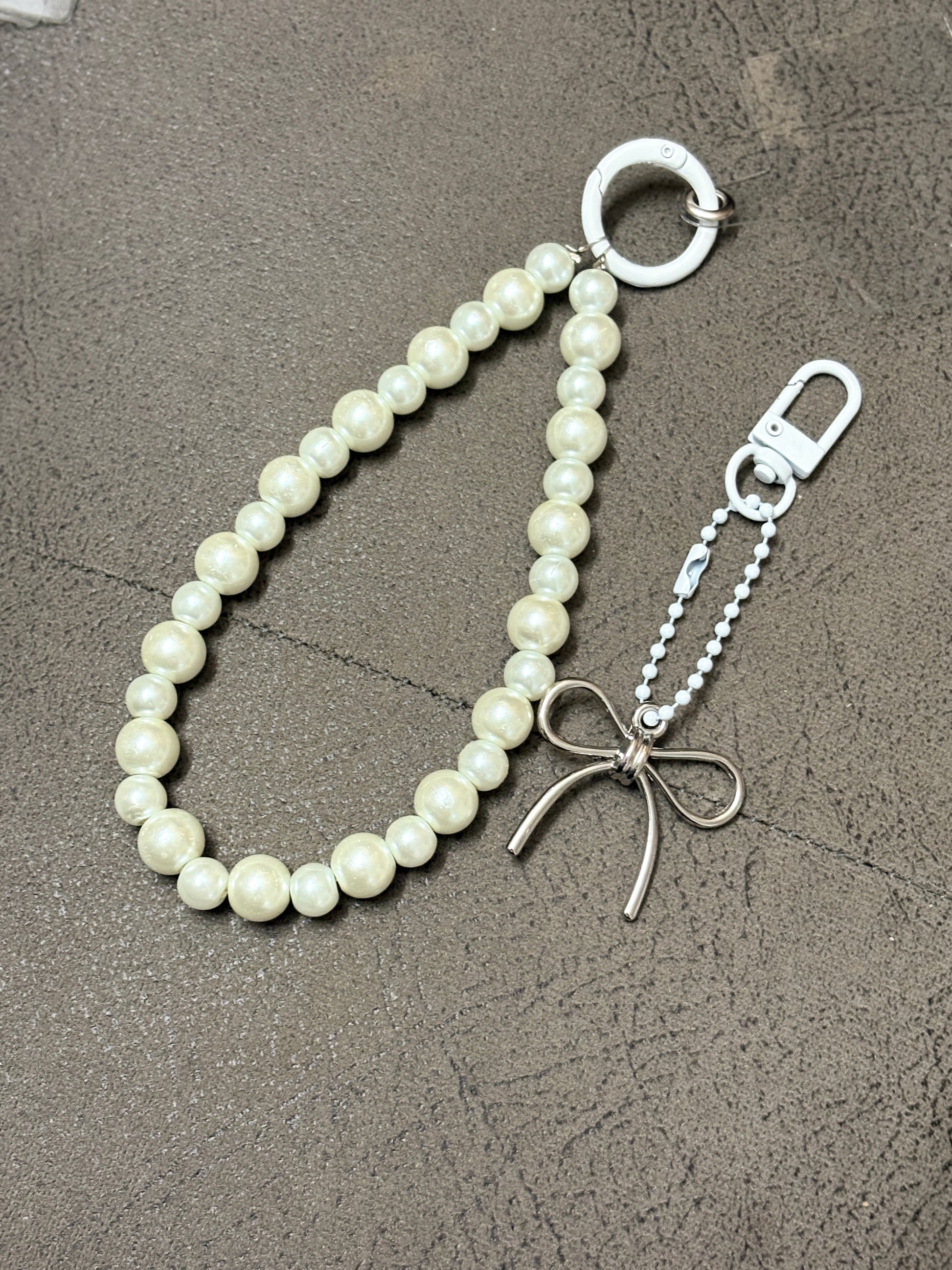 Elegant phone charm made with glossy pearl-like beads and a minimalist metallic bow charm, displayed on a textured background.
