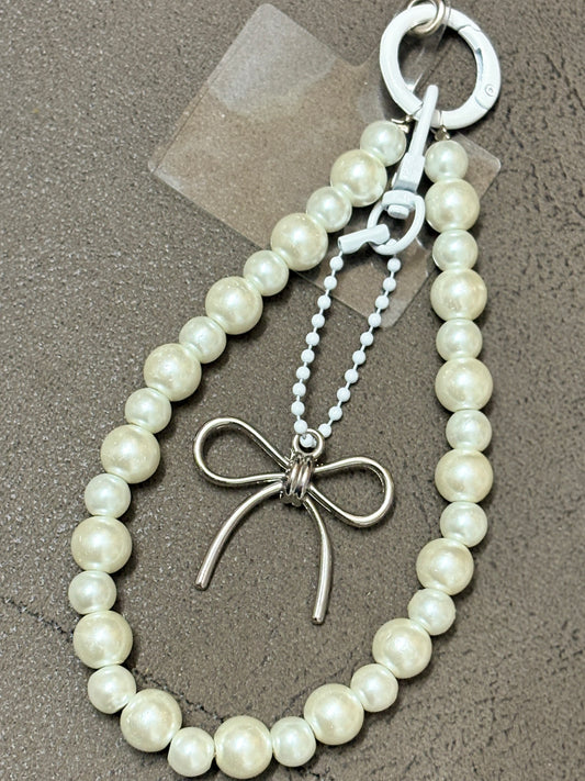 Elegant phone charm made with glossy pearl-like beads and a minimalist metallic bow charm, displayed on a textured background.
